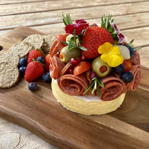 Baby Brie Cake piled high with charcuterie, olives and flowers on top.